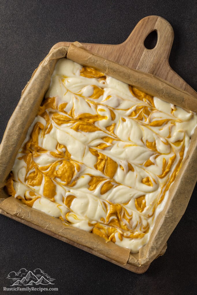 Swirled pumpkin and plain cheesecake batters over top of a crust in a baking pan.