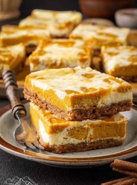 Two pumpkin cheesecake bars stacked on a plate next to a fork, with more cheesecake bars in the background.