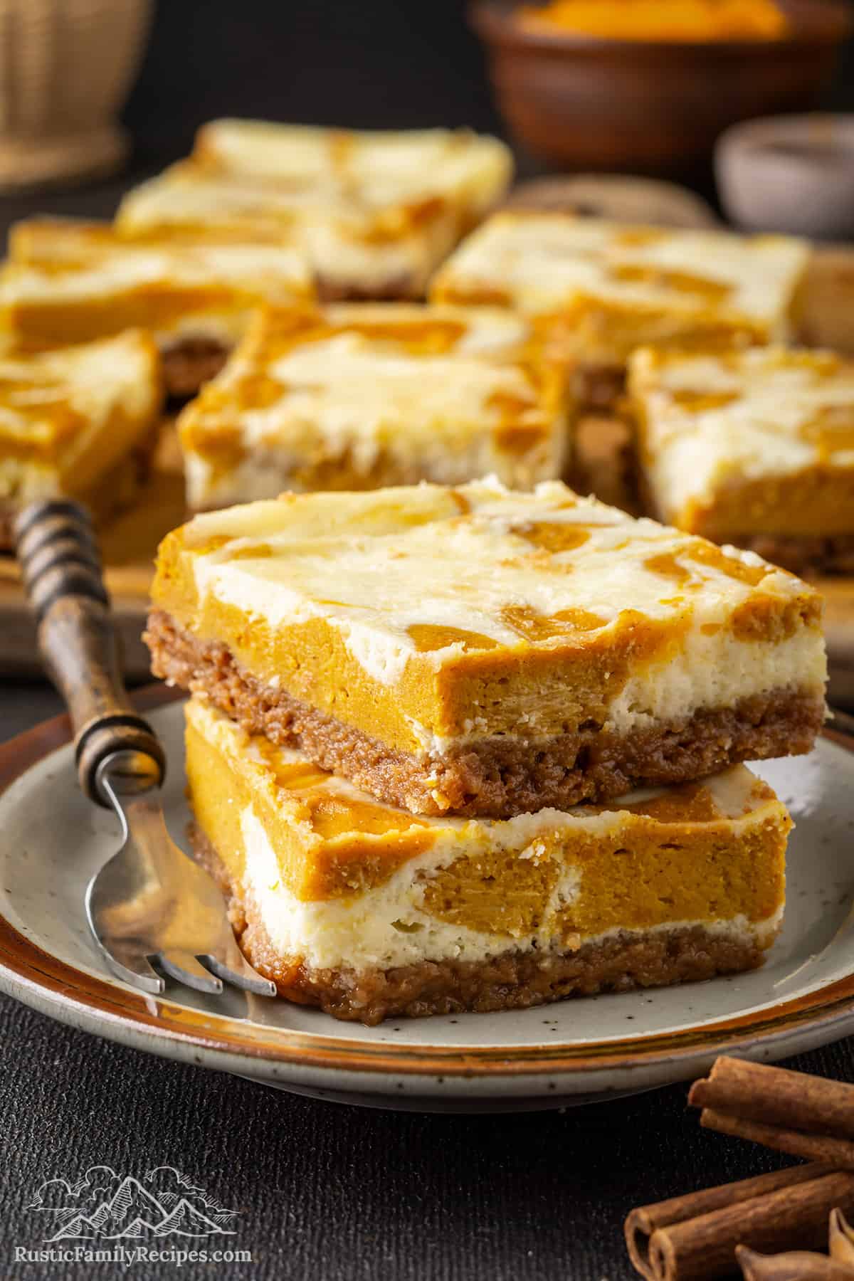 Two pumpkin cheesecake bars stacked on a plate next to a fork, with more cheesecake bars in the background.