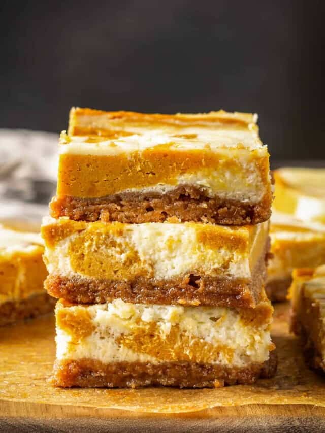 Three pumpkin cheesecake bars stacked on a wooden cutting board, with more bars in the background.