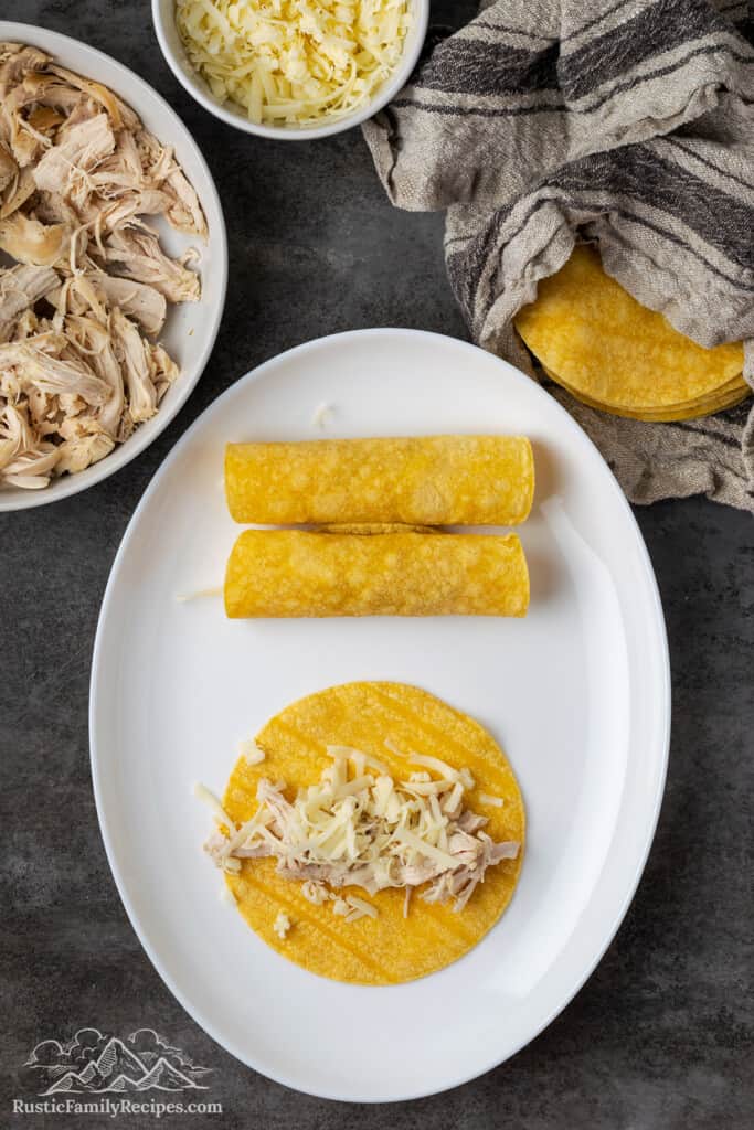 Shredded chicken added to the center of a corn tortilla on a plate, next to two rolled-up tortillas filled with chicken, surrounded by ingredients.