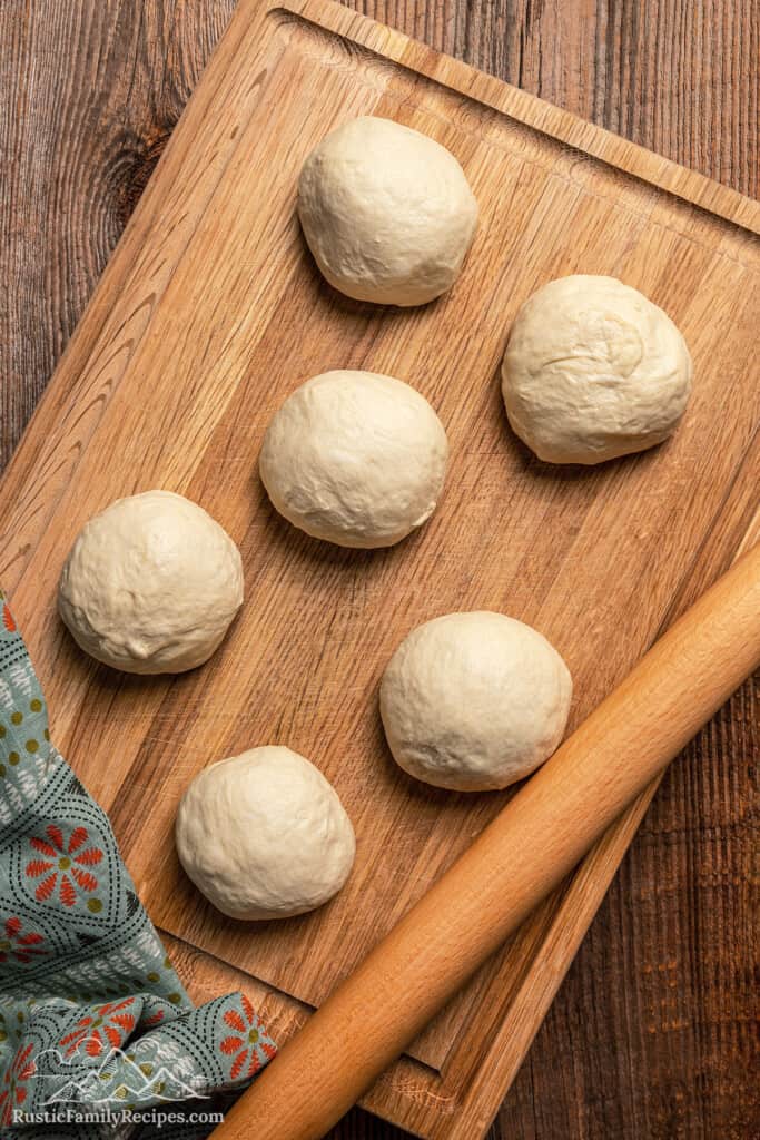 Six pizza dough balls portioned on a wooden cutting board next to a rolling pin.