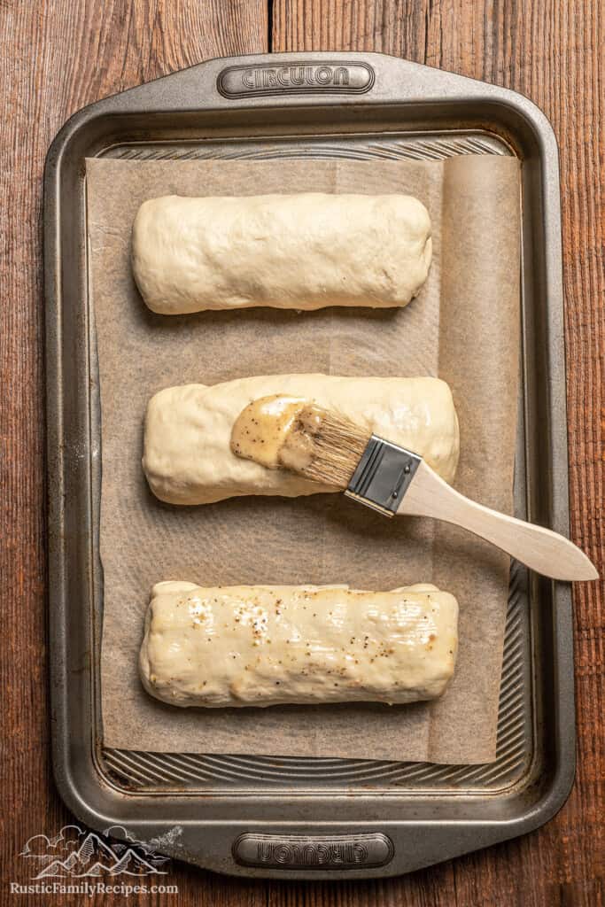 Three rolled chicken bakes are brushed with Caesar dressing on a baking sheet.