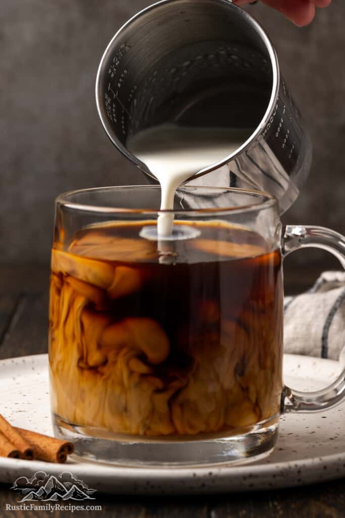 Pouring hot milk into a mug with coffee and cinnamon syrup.